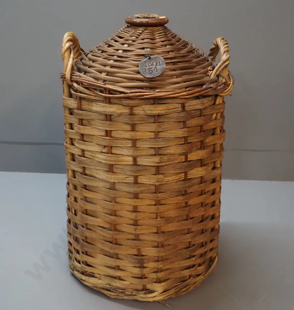 narrow-mouth stoneware crock in cane basket cover, with cork stopper and metal tag stamped 'M&H B54' H550 Image 1++