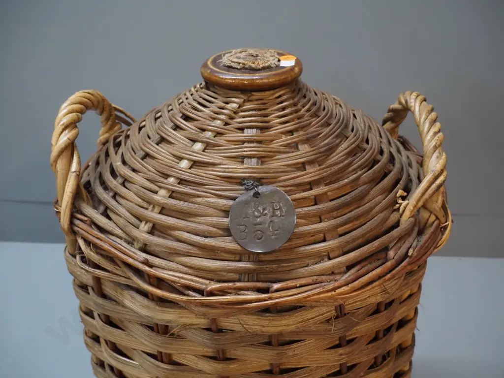 narrow-mouth stoneware crock in cane basket cover, with cork stopper and metal tag stamped 'M&H B54' H550 Image 1++
