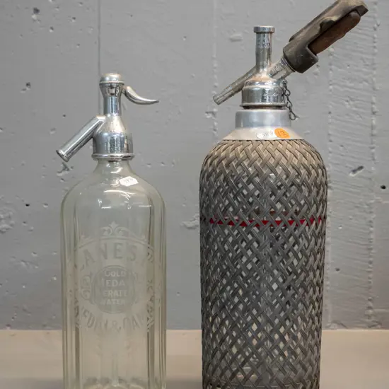 2 vintage soda syphons - one with Lane's Dunedin Oamaru mark to bottle and chrome top, other with wire mesh - some