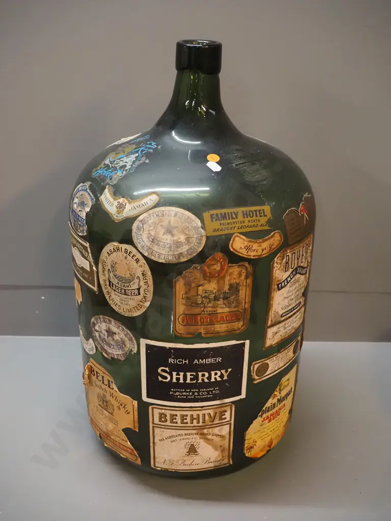 large dark green glass carboy covered in assorted labels including Family Hotel, Palmerston North, various drink Image 1++