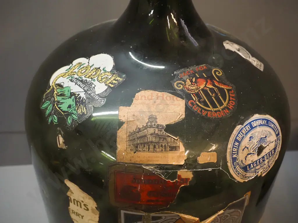 large dark green glass carboy covered in assorted labels including Family Hotel, Palmerston North, various drink Image 1++