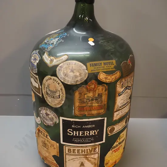 large dark green glass carboy covered in assorted labels including Family Hotel, Palmerston North, various drink