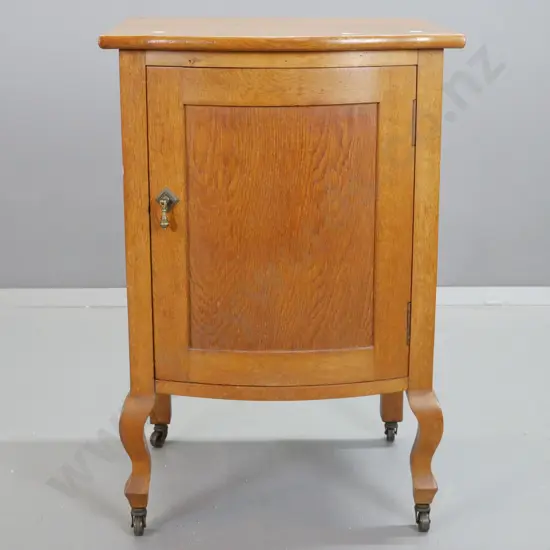 deco oak bow-front bedside cabinet with panelled door and sides, on short cab legs & metal casters, brass drop handle.