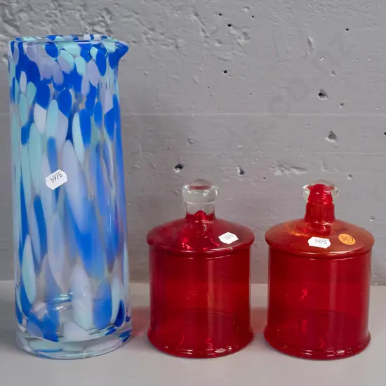 3 glass items - 2 red lidded canisters, one with clear knob, and a blue mottled art glass tall vase or jug with pouring
